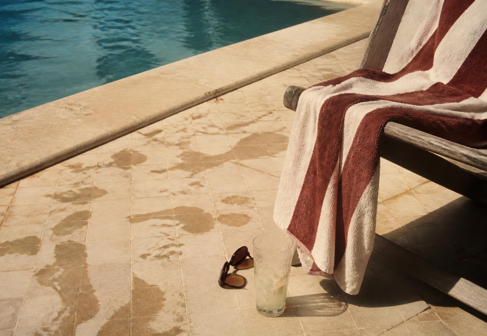 A damp striped pool towel over a chair beside sunglasses and an unfinished Paloma